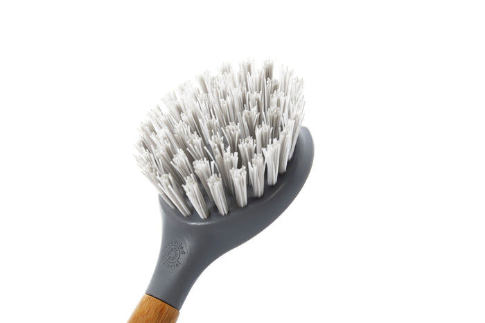 A close-up of the Full Circle Tenacious C Cast Iron Brush features stiff, recycled-nylon bristles and a sleek grey base. A wooden handle partially visible at the bottom adds a classic touch, all set against a plain white background.