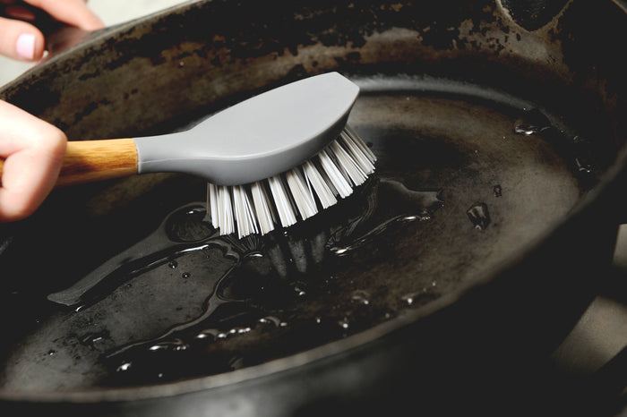A hand scrubs a greasy, blackened skillet with the Full Circle Tenacious C Cast Iron Brush, featuring a bamboo handle and recycled-nylon bristles. Water shimmers on the pans surface as residue is scrubbed away.