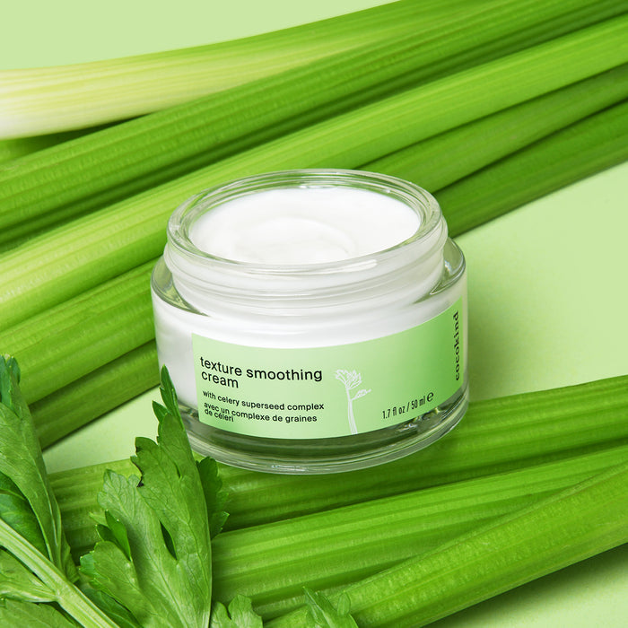 A jar of Cocokinds Texture Smoothing Cream, featuring Celery Superseed Complex and Tsubaki seed oil, is displayed on a green surface surrounded by fresh celery stalks and leaves. The open jar reveals the smooth white cream inside.