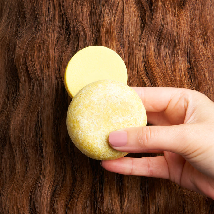 A hand holds two round, yellow bars from The Balancer Travel Set by NOT!CE Hair Co against wavy, reddish-brown hair. The soft pink nails highlight a pledge to plastic-free hair care.