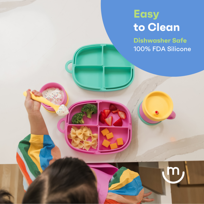 A child wearing a colorful striped outfit is sitting at a table with a pink and green divided silicone plate containing pasta, broccoli, strawberries, and cheese cubes. A matching green lidded cup is nearby. Text reads "Easy to Clean, Dishwasher Safe, 100% FDA Silicone.