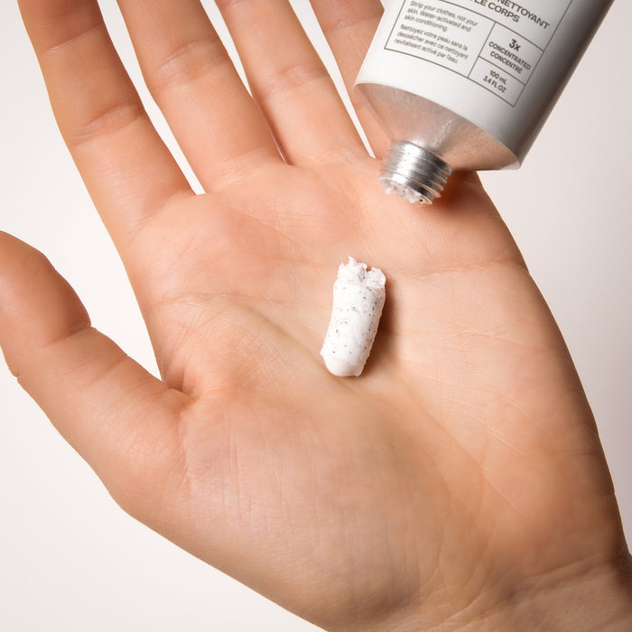 A hand holds a small amount of white, textured The Exfoliating Body Wash Concentrate by Everist, squeezed from a silver tube and resting on the open palm against a plain background.