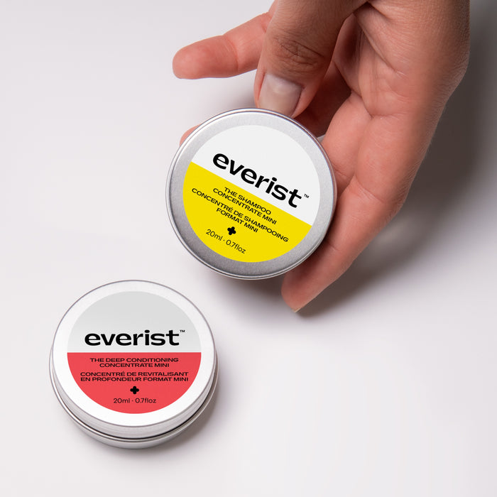 A hand holds Everists The Haircare Travel Duo—The Shampoo Concentrate with a yellow stripe and The Deep Conditioning Concentrate with a red stripe—spotlighting waterless, plastic-free haircare on a white surface.