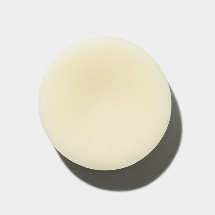 A round, pale yellow object resembling a button or soap is centered against a white background with its shadow beneath it. The Hydrator Conditioner Bar from NOT!CE Hair Cos plastic-free line promises to moisturize hair with every use.