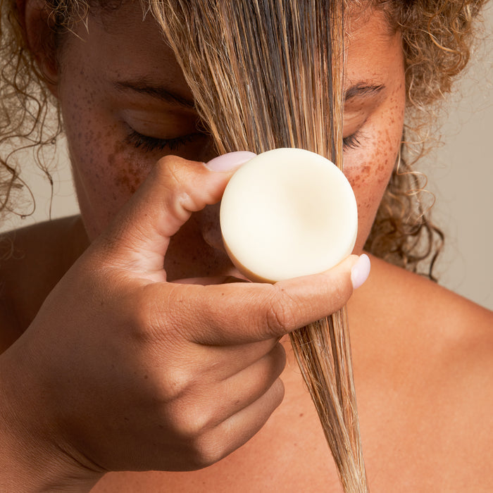 A curly-haired person uses The Hydrator Conditioner Bar by NOT!CE Hair Co, a plastic-free solution that glides through damp hair. Infused with essential oils, it nourishes and revitalizes curls while moisturizing.