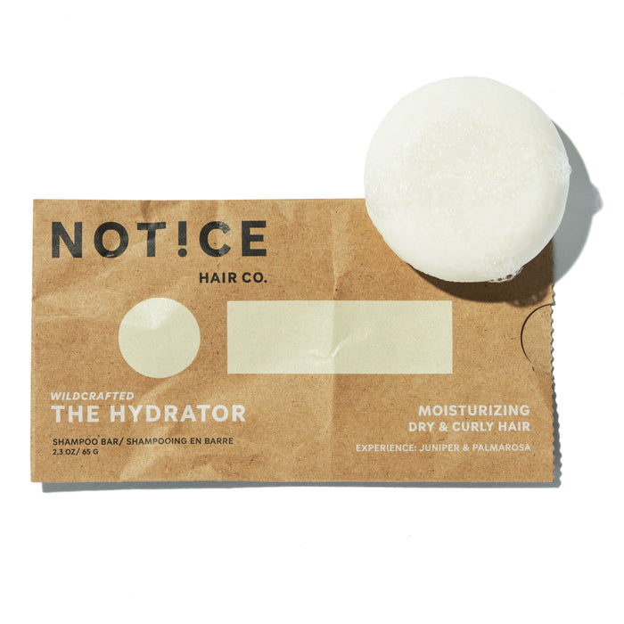 A circular white shampoo bar rests on brown packaging labeled NOT!CE Hair Co. The Hydrator Shampoo Bar. It offers moisturizing care for dry, curly hair with juniper and palmarosa experiences. Enjoy this plastic-free solution.