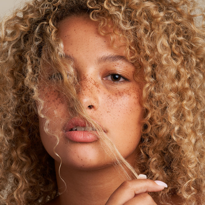 A person with curly blonde hair and freckles holds a strand of their hair near their face, gazing directly into the camera against a neutral background, highlighting the benefits of using The Hydrator Shampoo Bar by NOT!CE Hair Co for moisturized locks.