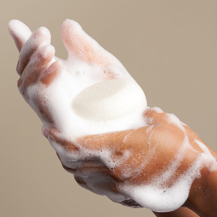 Hands covered in white foam hold The Hydrator Shampoo Bar by NOT!CE Hair Co against a beige background, highlighting a dedication to plastic-free hair care.