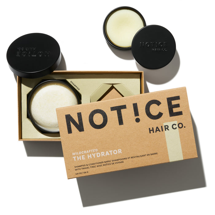 An open brown box labeled NOT!CE Hair Co reveals The Hydrator Travel Set, containing a jar of cream and two black lids with NOT!CE text. This plastic-free hair care set, featuring an innovative hydrator shampoo, is arranged on a light surface with shadows.
