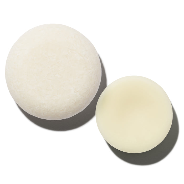 The Hydrator Travel Set by NOT!CE Hair Co features two round off-white solids on a white backdrop. One is textured, resembling a detailed conditioner bar, and the other is smooth like a hydrator shampoo. Both cast delicate shadows, showcasing plastic-free hair care aesthetics.