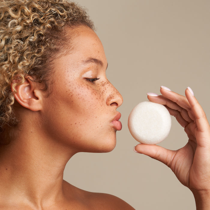 With curly hair and freckles, a person holds a round, white object near their lips, gazing at it. In the spirit of plastic-free hair care, perhaps its from The Hydrator Travel Set by NOT!CE Hair Co. The background is neutral.