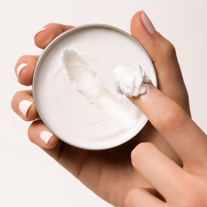 A hand with light pink polished nails holds an open tin of Everist The Shampoo Concentrate Tin. A finger scoops out the thick, smooth formula, enriched with natural rosemary oil, against a plain light background.