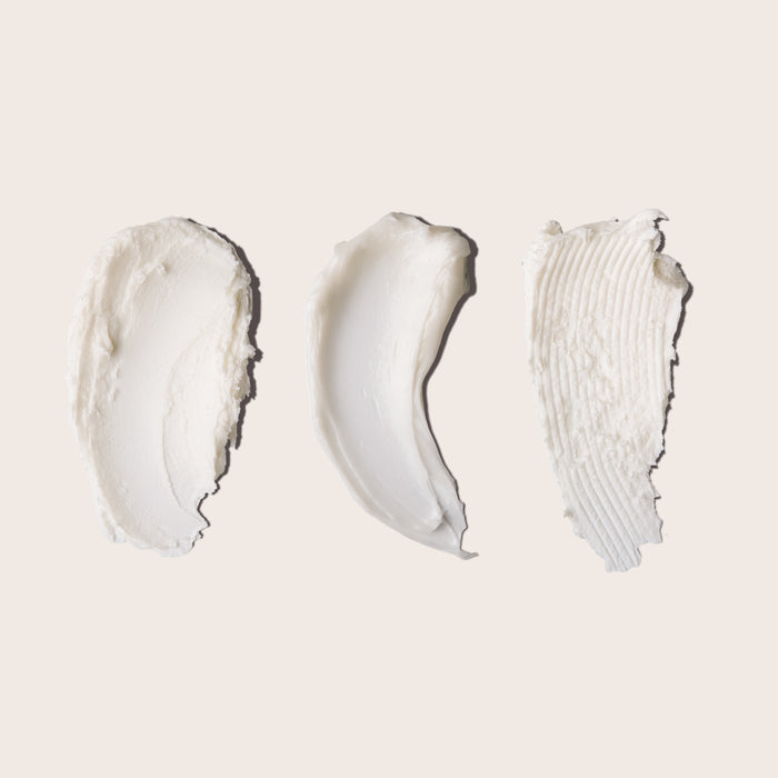 Three swipes of white creamy formulas—each with a unique texture and pattern—reminiscent of Everists The Shower Essentials Travel Kit products, are displayed on a light beige background.