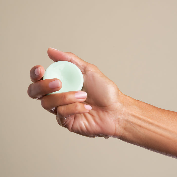 A hand presents The Stimulator Conditioner Bar by NOT!CE Hair Co, a small, round, light green bar against a plain beige backdrop. Slightly wet, it highlights this innovative, plastic-free hair care solution crafted to nurture and boost hair growth.