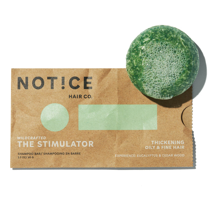 A green shampoo bar with foam sits on a brown paper package labeled NOT!CE Hair Co. and The Stimulator Shampoo Bar. This plastic-free option is designed to thicken oily and fine hair, infused with eucalyptus and cedar wood for promoting hair growth.