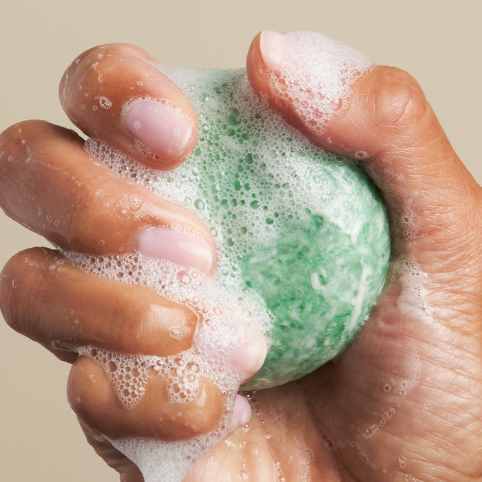 A hand with neatly manicured, pink-tinted nails tightly squeezes The Stimulator Shampoo Bar by NOT!CE Hair Co, creating a lather of white suds. This plastic-free hair care choice stands out against the neutral beige background.