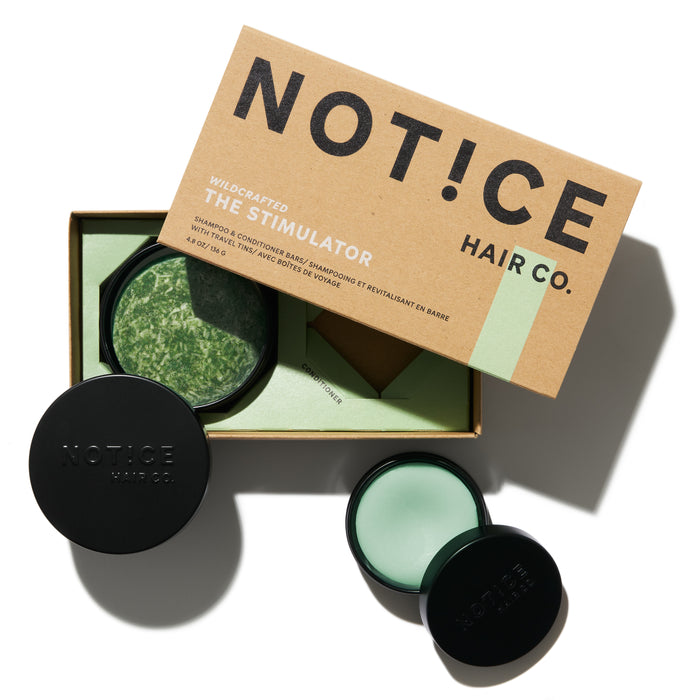 A cardboard box labeled NOT!CE Hair Co houses The Stimulator Travel Set next to round green and black containers with French green clay, featuring the brand name on the lid, hinting at premium shampoo and conditioner bars.