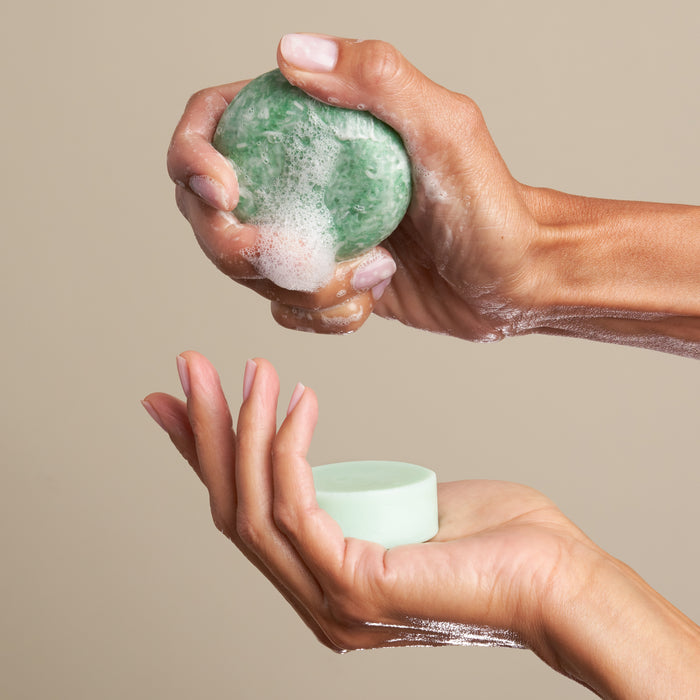 Two hands are seen, one squeezing The Stimulator Travel Set by NOT!CE Hair Co. Its a green, foam-covered soap with French green clay. An open hand is beneath it against a neutral beige background.