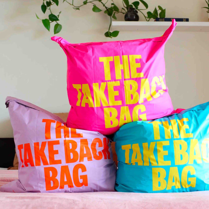 Stacked together, Trashies The Take Back Bag features three colorful bags: pink with yellow text, lavender with orange text, and blue with yellow text. Ideal for collecting unwanted clothes, a plant on a shelf adds ambiance in the background.