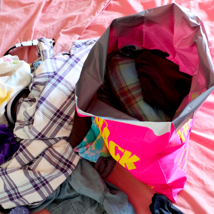 A Trashie pink & black Take Back Bag brims with vibrant clothes on a bed. Garments in plaid and solid styles are artfully laid out next to the bag on a pink sheet.