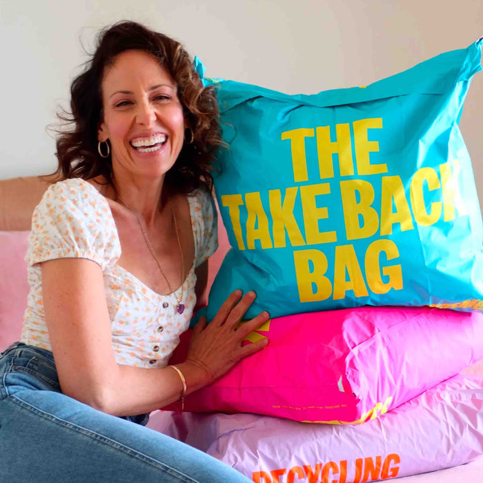 A woman beams, leaning on colorful bags like Trashies The Take Back Bag for unwanted clothing. Dressed in a polka dot blouse and jeans, the rooms cheerful pink and blue hues create an ideal setting for the TrashieCash™ revolution.
