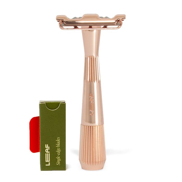 A rose gold Twig Plastic-Free Razor from Leaf Shave, featuring an ergonomic handle, stands upright against a white background next to a small green box labeled LEAF containing single-edge blades.