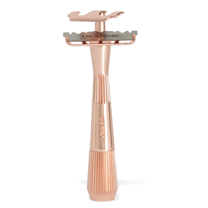 The Twig Plastic-Free Razor by Leaf Shave is a rose gold metal safety razor with a single-edge blade, featuring an ergonomic, textured handle with vertical ridges for a superior grip. It stands upright and boldly declares against plastic use.