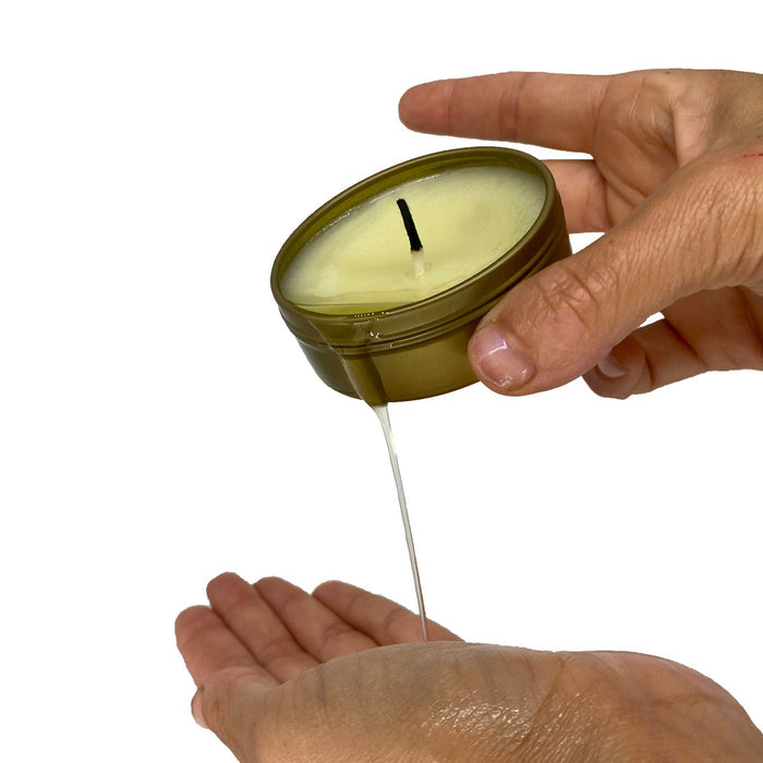 Hands cradle a small metal tin of the ThreePlay Massage Candle by Lovability, pouring the melted wax onto an open palm. This 3-in-1 candle soothes and ultra-hydrates, solidifying around its wick, with a minimalistic white background highlighting its elegance.