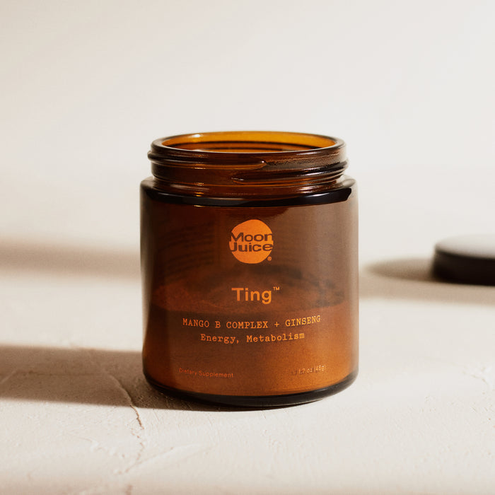 A brown Moon Juice jar labeled Ting - Mango B Complex + Ginseng for Energy & Metabolism is open, with the lid placed next to it on a light-textured surface.