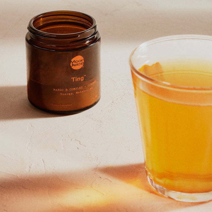 A glass of orange liquid on a light surface is next to a brown jar labeled Ting, containing Moon Juices Mango B Complex + Ginseng for Energy & Metabolism. Warm natural light bathes the scene, embodying wellness and vitality.