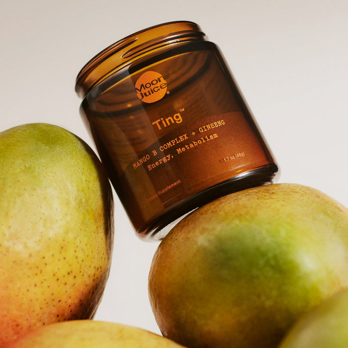 A Moon Juice jar labeled Ting - Mango B Complex + Ginseng for Energy & Metabolism is balanced on fresh mangoes, featuring an amber-colored round design.