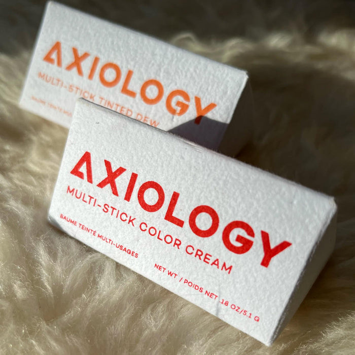Two Axiology product boxes, one labeled Tinted Dew and the other Multi-Stick Color Cream, sit on a soft surface. These multi-use color items feature bold red text on compostable packaging, emphasizing sustainability.