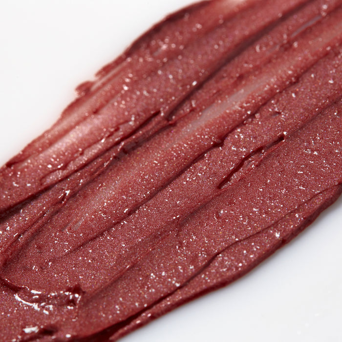The sentence can be rewritten as follows: A close-up of Tinted Dew by Axiology reveals a smooth smear of shimmery, metallic reddish-brown makeup against a white background. Its glossy texture with sparkles creates a lustrous look. This versatile color comes in compostable packaging, making it stunning and eco-friendly.