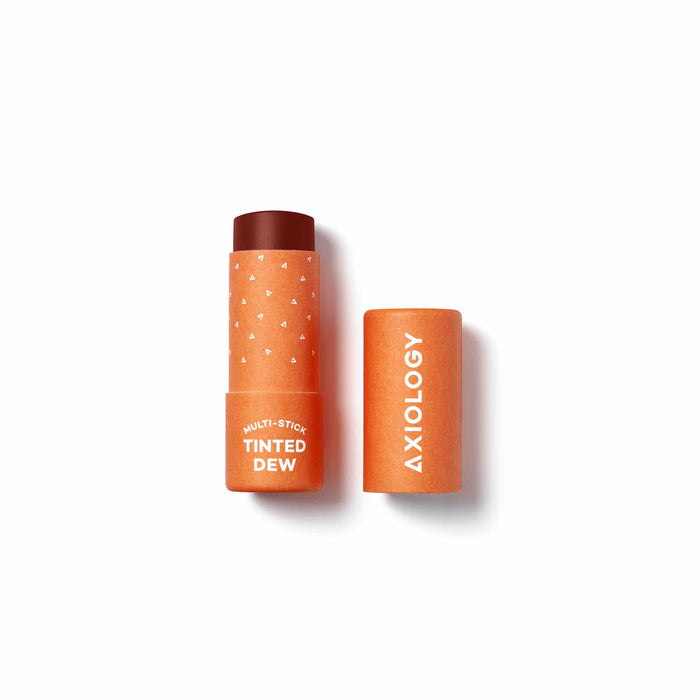The Axiology Tinted Dew multi-stick, in its orange tube with the lid off, reveals the product. The eco-friendly packaging features small white heart patterns and the brand name in white text, crafted from compostable materials.
