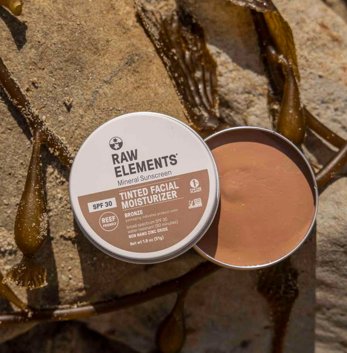 An open Tinted Tin Bronze by Raw Elements displays its bronze ultra-moisturizing cream. Resting on sand with seaweed, the recyclable tin highlights its reef-safe, natural ingredients.
