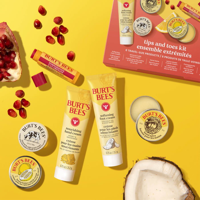 Flat lay of Burts Bees Tips & Toes Set, featuring hand creams, balms, and salves, with pomegranate seeds—highlighting the Pomegranate Lip Balm—and coconut halves on a yellow background. The set’s box is visible in the upper right corner.