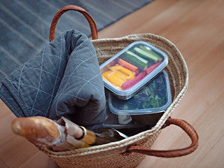 A woven basket on the floor contains a quilted gray blanket, a baguette, a bottle of wine, and eco-friendly U-Konserve To-Go Large Containers packed with an assortment of cut vegetables and leafy greens.