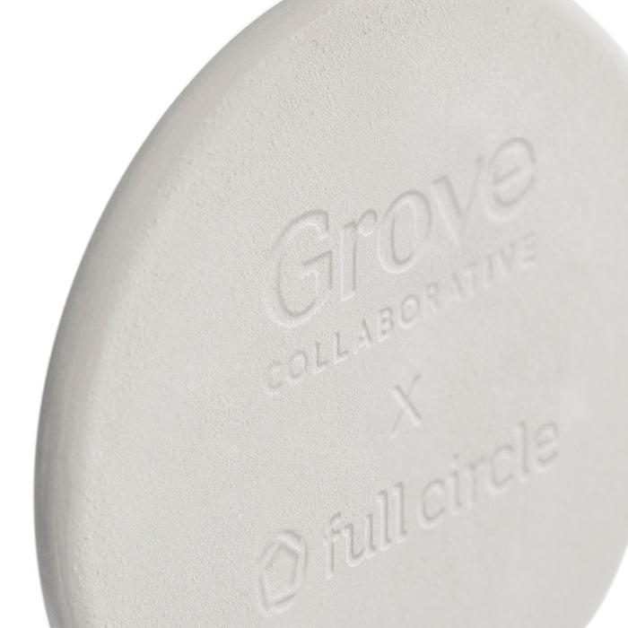 Close-up of a round, light grey Toilet Brush Drip Stone Refill by Grove Co., made from all-natural diatomaceous earth. It features a smooth, matte texture and is embossed with Grove Collaborative X Full Circle, designed to prevent bacteria and mold growth.