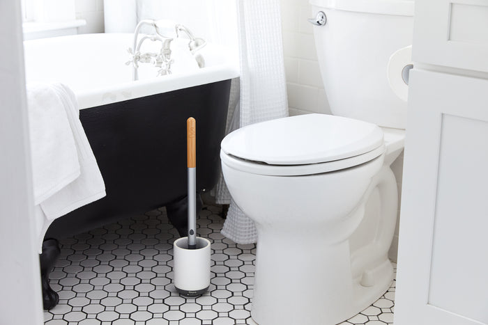 A clean, white bathroom showcases a classic bathtub with a dark exterior and silver fixtures. Nearby sits a white toilet, accompanied by a Grove Co. Toilet Brush with Replaceable Head resting in its holder on the hexagonal-tiled floor. White towels hang nearby.