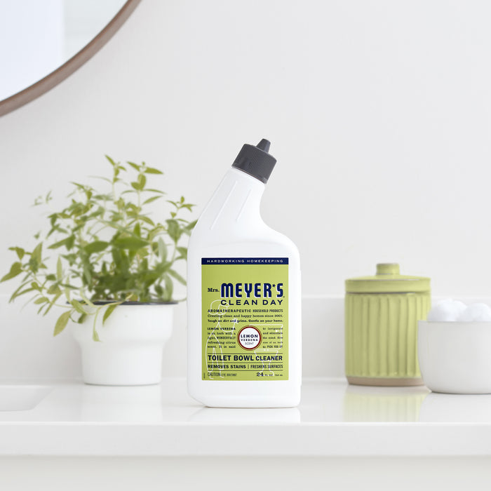 A Mrs. Meyers Toilet Cleaner bottle sits on a white bathroom countertop, with a small potted plant and green jar visible in the background.