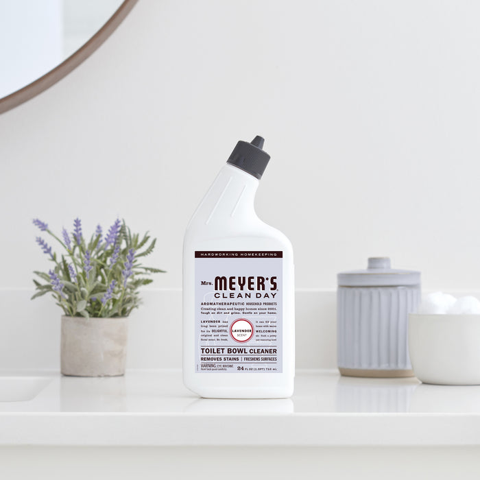 A bottle of Mrs. Meyers Toilet Cleaner with essential oils and a dark nozzle is on a white bathroom counter, while a plant and cotton balls container enhance the labels lavender scent focus and cleaning benefits.