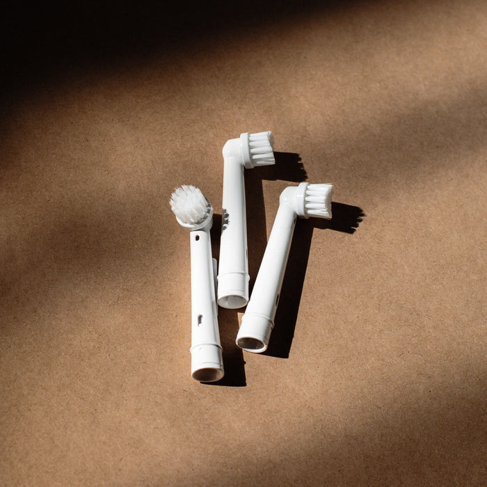 Three Plus Ultra eco-friendly Toothbrush Head Replacements compatible with Oral-B lie on a brown surface, softly lit, casting gentle shadows. Their bristles vary in shape and length for a perfect fit.