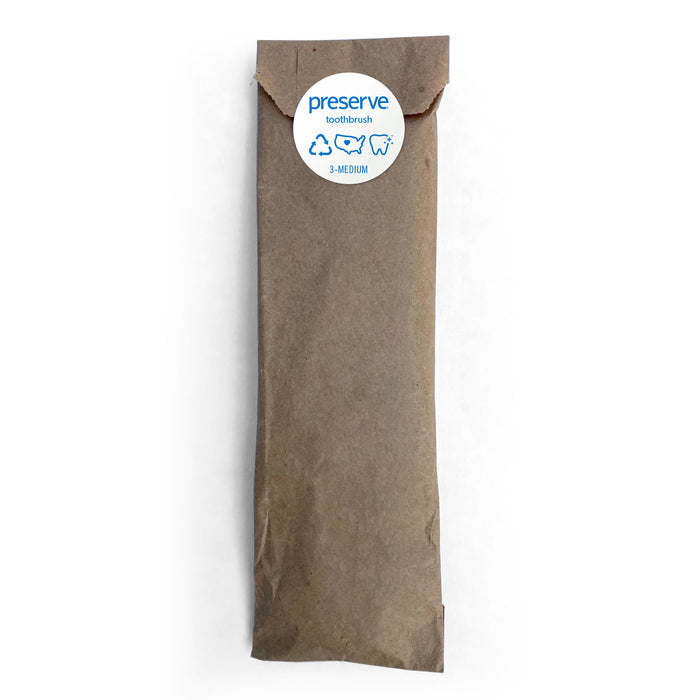 A brown paper pouch with a round white sticker and blue text contains a Preserve toothbrush made from recycled plastic, labeled Preserve Toothbrush (Soft), and features recycling symbols to emphasize its eco-friendly qualities.