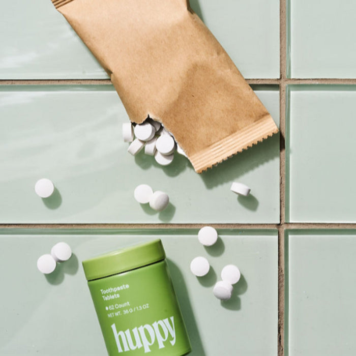 A brown paper pouch of Huppy Toothpaste Tablets Refill spills white tablets onto light green tiles. Next to it stands a green Huppy container labeled Toothpaste Tablets—your sustainable toothpaste for cavity fighting.