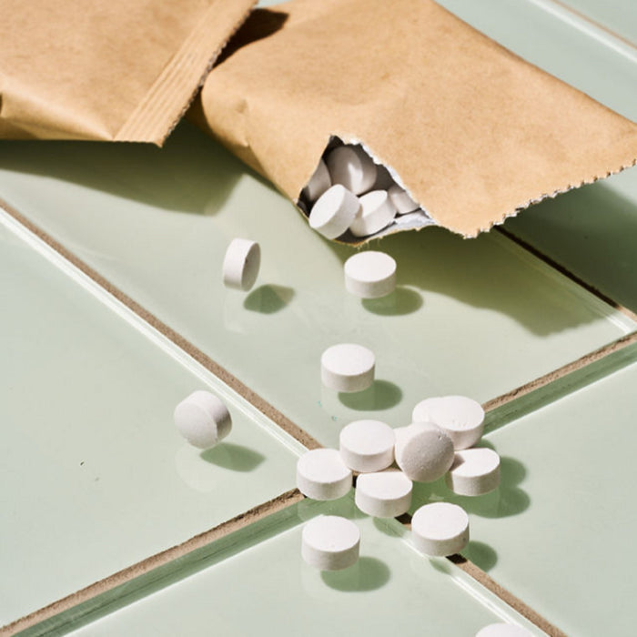 A brown paper pouch of Huppy Toothpaste Tablets Refill spills several round white tablets onto a shiny, pale green tiled surface—an eco-friendly, sustainable alternative for effective cavity care.