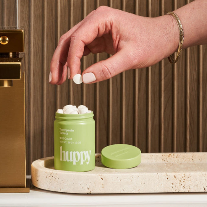 A hand holds a Huppy Toothpaste Tablets Refill above an open green jar filled with white tablets for natural whitening, resting on a beige tray beside a gold sink fixture.