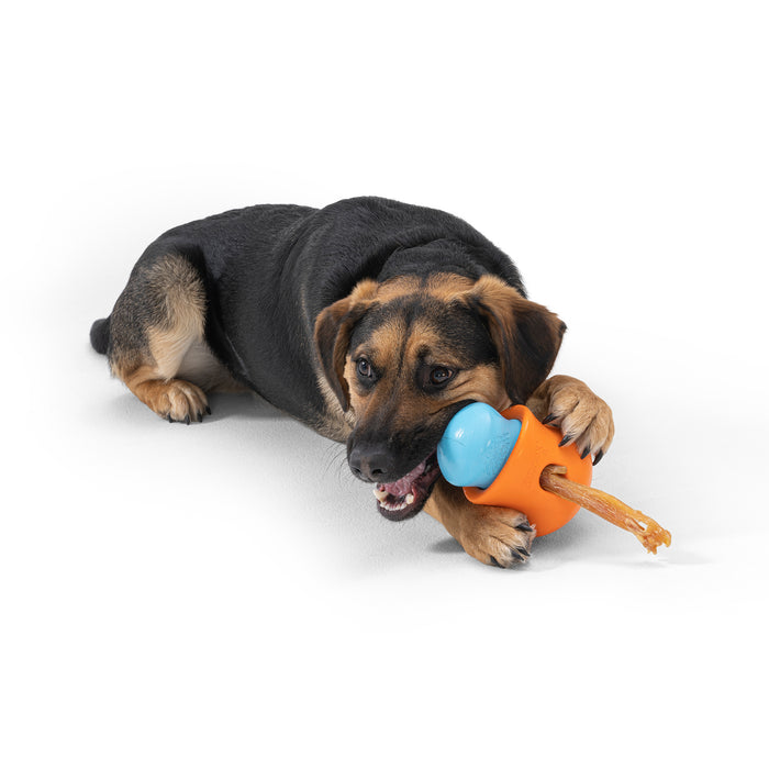 A brown and black dog is happily lying down, chewing on a West Paw Treat Dispenser Dog Toy. The toys playful challenge keeps the pup engaged against a plain white background.