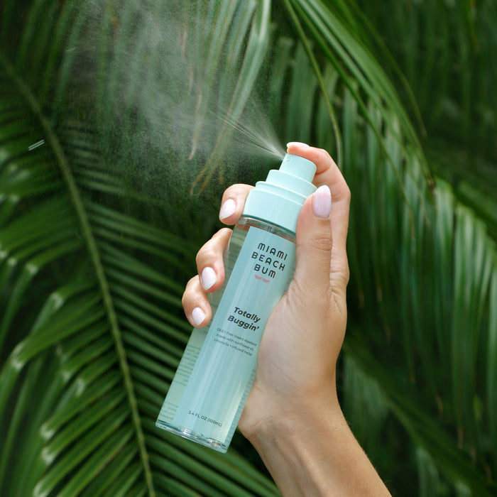 A hand holds a Totally Buggin spray by Miami Beach Bum in front of lush green palm leaves. This DEET-free, natural insect repellent releases a gentle mist with its pH-balanced formula, ready to protect.
