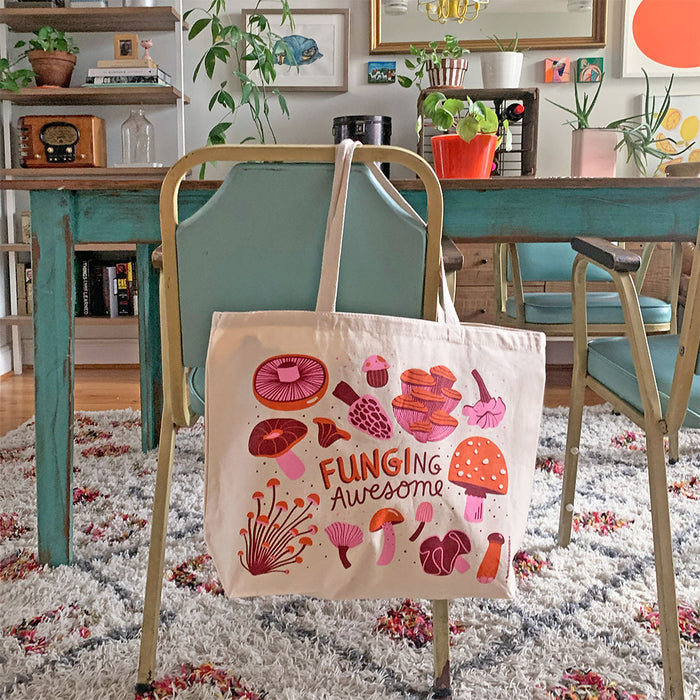 A The Neighborgoods cotton tote bag with vibrant mushroom designs and Funging Awesome text hangs on a chair. Crafted from durable canvas, it sits by a dining table amid plants, eclectic décor, and a patterned rug—perfect as an eco-friendly gift bag.