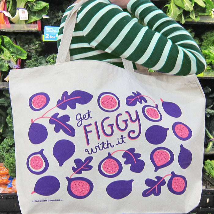 A person in a green and white striped shirt holds The Neighborgoods Tote Bag featuring a colorful fig design and the text get figgy with it. Shelves filled with leafy greens and fresh produce are visible in the background.